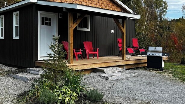 Een klein vakantiehuisje met een veranda, rode stoelen en een barbecue.