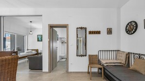 Een moderne hotelkamer met een bed, een bank, een stoel en een grote spiegel. Een gang leidt naar een andere kamer met een bed en een raam.