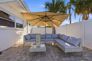 A sectional sofa with a glass-top coffee table and a beige umbrella in a backyard patio area.