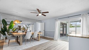 Un espace de salle à manger moderne, doté d’un ventilateur de plafond, d’une table à manger avec des chaises, d’une grande plante en pot et d’une vue sur l’extérieur, accessible par une porte-fenêtre coulissante.