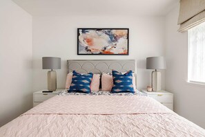 A bedroom with a bed, two bedside lamps, a nightstand, a wall-mounted abstract painting, and a window with curtains.