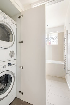 A laundry room with a front-loading washer and dryer, a white door, and a view into a bathroom with a bathtub and a window.