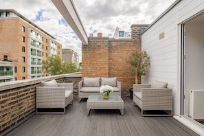 A balcony with wicker furniture, a glass table, and a brick wall.