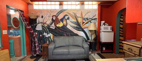 Un salon chaleureux avec un canapé gris, une fresque murale colorée et un petit coin cuisine équipé d’un four à micro-ondes.
