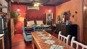 Une pièce de taille modeste, avec une table à manger carrelée, un lit recouvert d’une couverture à rayures, un petit chien blanc et un ventilateur de plafond.