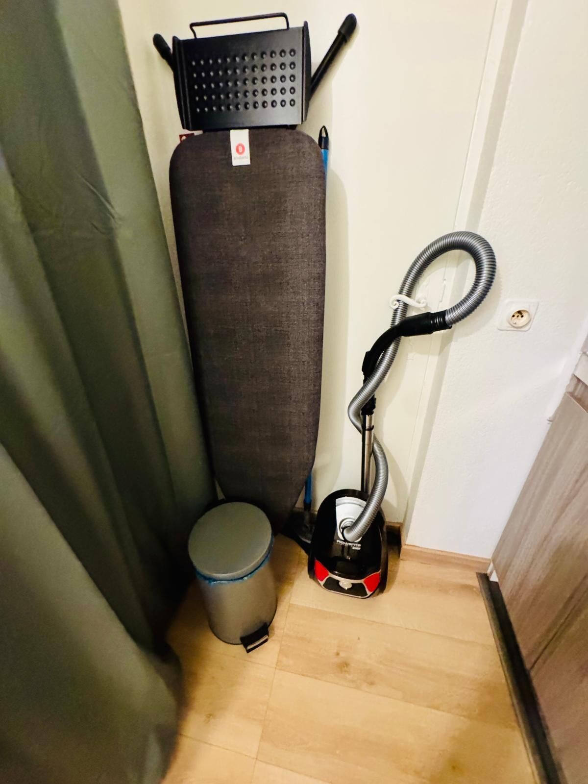 Une petite pièce étroite, avec un canapé gris, un aspirateur, une poubelle et une prise de courant au mur.