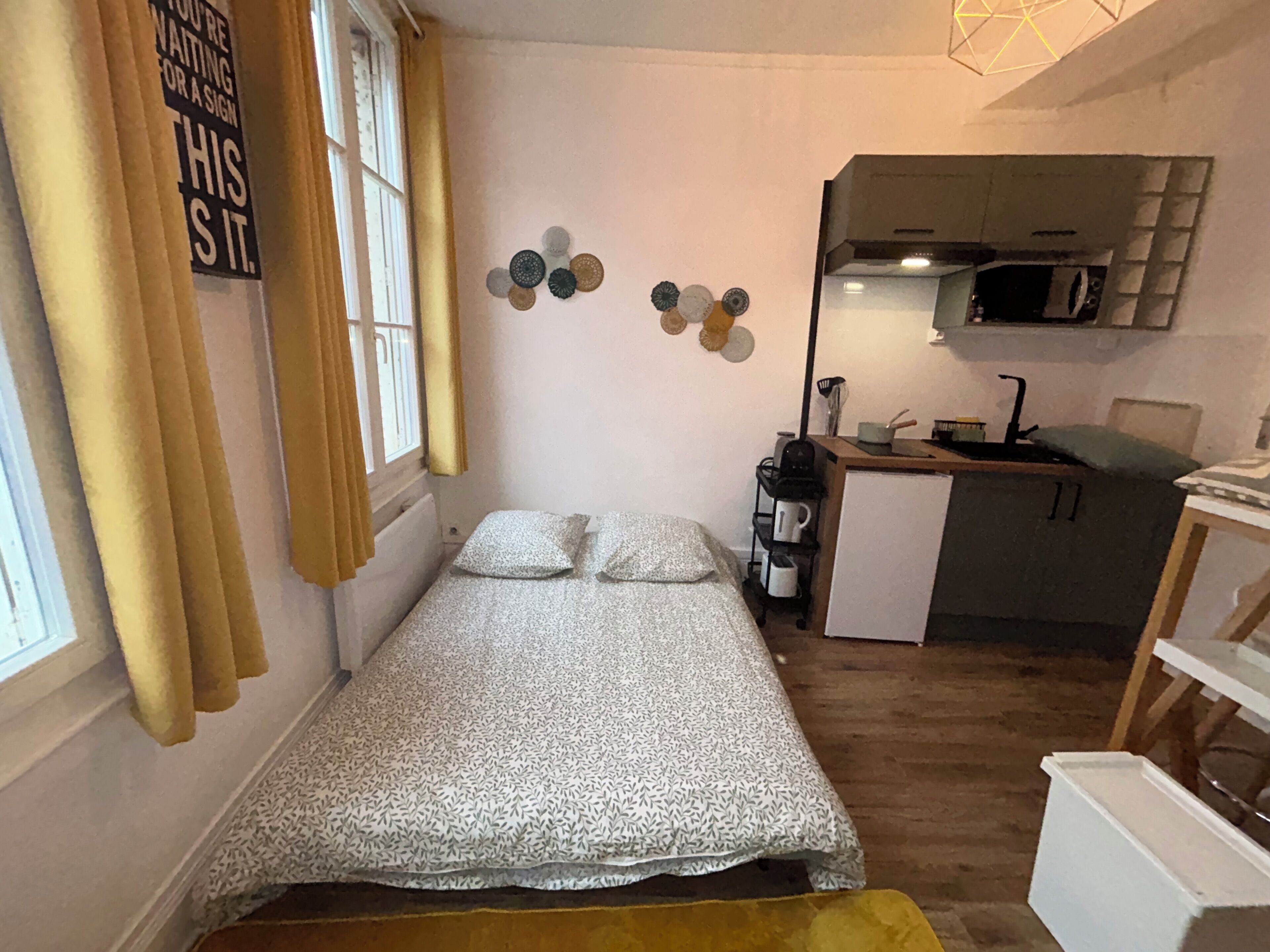 A small, compact room with a bed, a kitchenette, and a window with curtains.