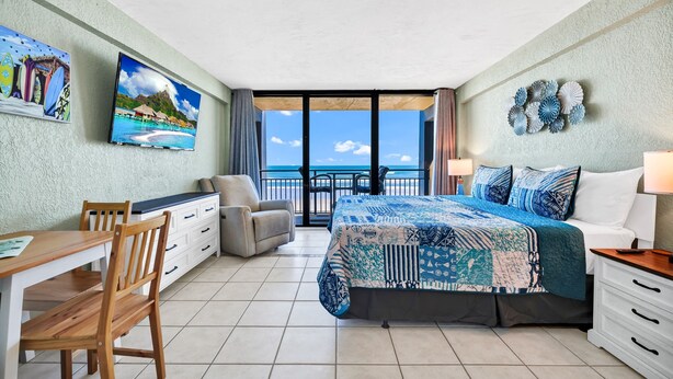 A bedroom with a bed, a desk, a chair, a TV, and a balcony with a view of the ocean. -  Studio for 2 with Breathtaking View with A/C (Daytona Beach Shores)
