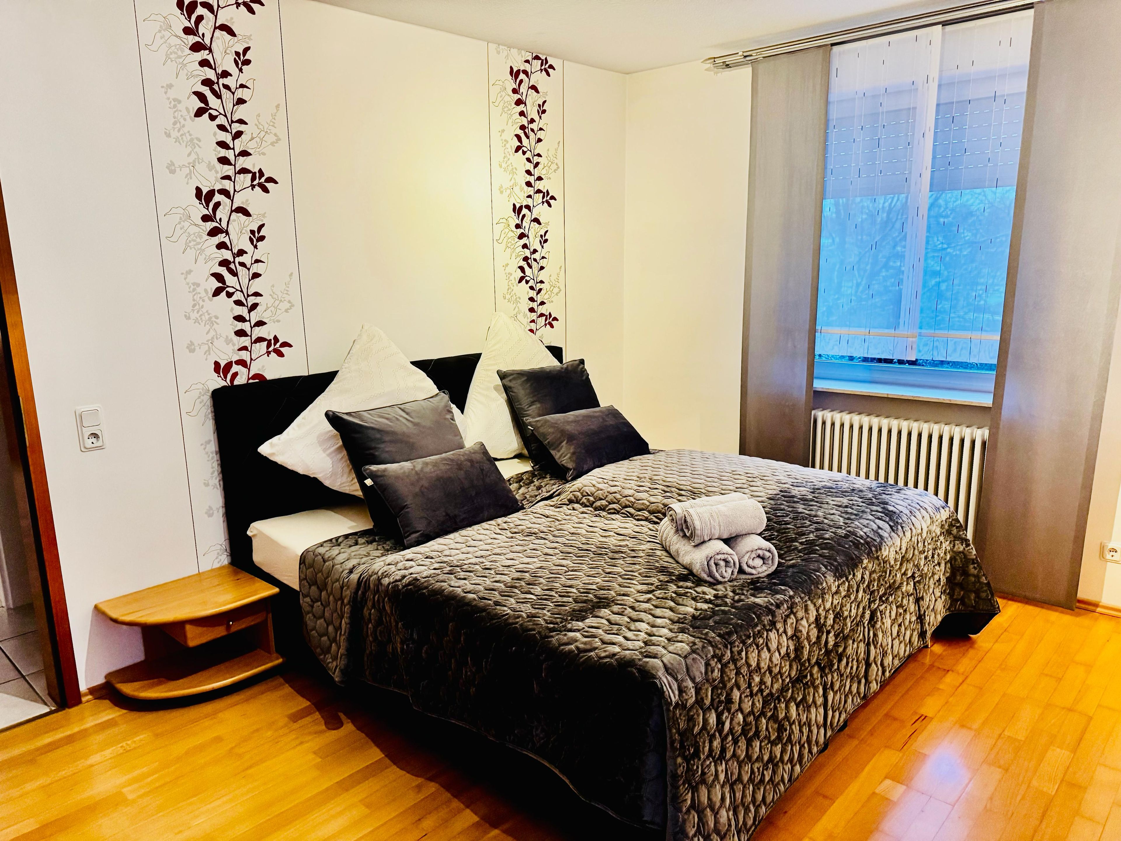 A bedroom with a bed, pillows, a bedside table, a radiator, and a window.