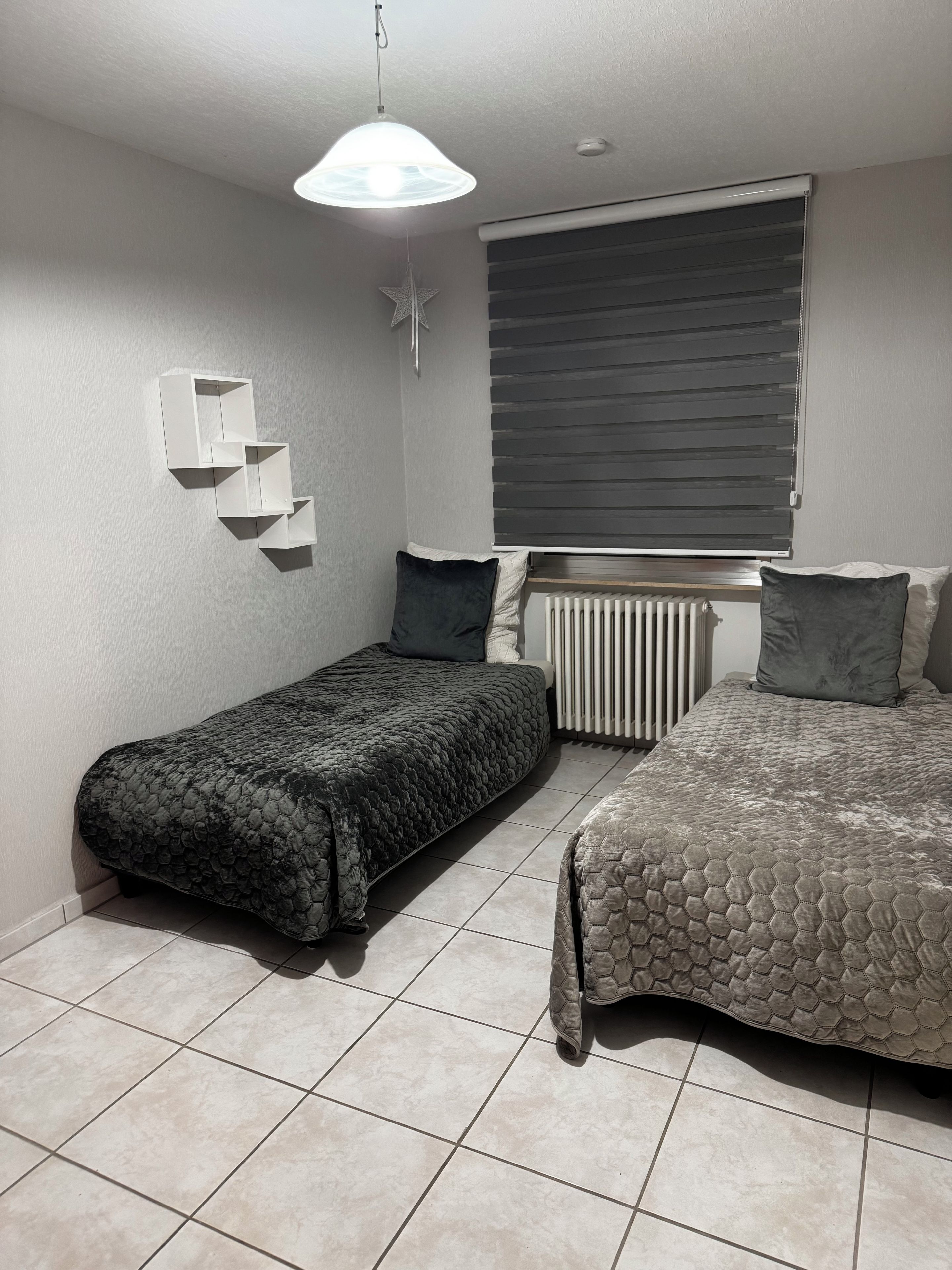 Ein Schlafzimmer mit zwei Betten, einem Heizkörper, einem Fenster mit Jalousien und einem Wandregal.