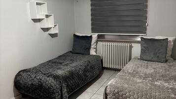 Ein Schlafzimmer mit zwei Betten, einem Heizkörper, einem Fenster mit Jalousien und einem Wandregal.