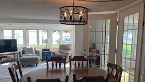 A dining area with a round table and chairs, a television, and a view of the outdoors through large windows. - Stay At The Lake House (Houghton Lake)