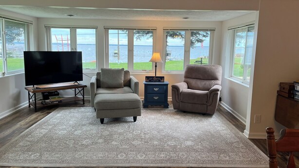A living room with a large window overlooking a body of water, a flat-screen TV, two armchairs, a small table with a lamp, and a rug. - Stay At The Lake House (Houghton Lake)