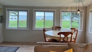 A dining area with a round table and chairs, large windows with a view of the sea, and a hanging light fixture. - Stay At The Lake Cottage (Houghton Lake)