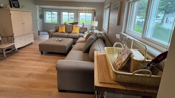 A spacious living room with a sectional sofa, ottoman, and a dining area with a table and chairs. - Stay At The Lake Cottage (Houghton Lake)