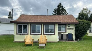 A small, single-story house with a brown roof and light green siding, featuring two wooden chairs and a small table in the front yard. - Stay At The Lake Cottage (Houghton Lake)