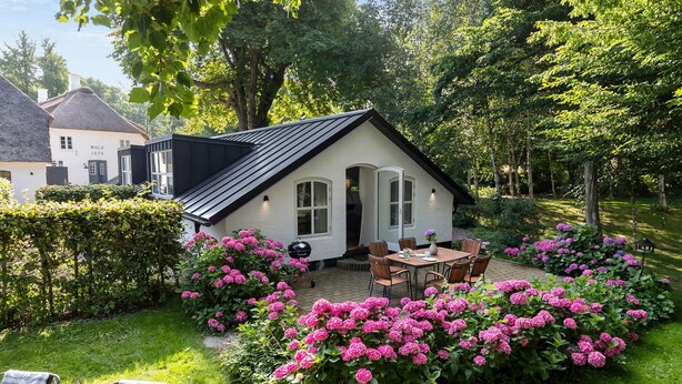 A small house with a thatched roof, surrounded by a garden with pink flowers and a patio with a table and chairs. - Beautiful home in Aabenraa with house sea view (Aabenraa)