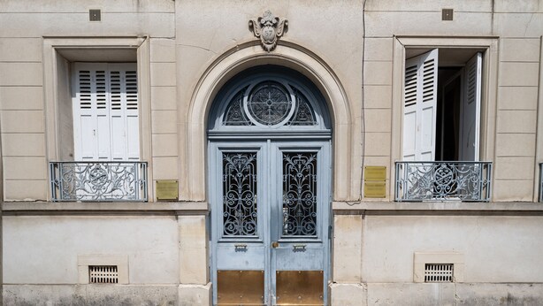 A building facade with a central arched doorway, flanked by two windows with shutters, and a decorative balcony railing.