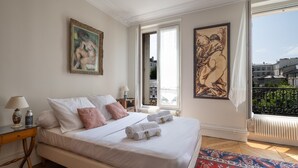 A hotel room with a large bed, a nightstand, a window with a view of buildings, and two paintings on the walls.