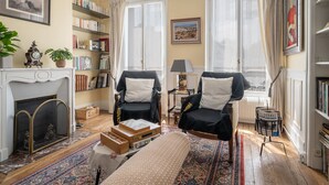 A cozy living room with a fireplace, two armchairs, a bench, a bookshelf, and a view through large windows.