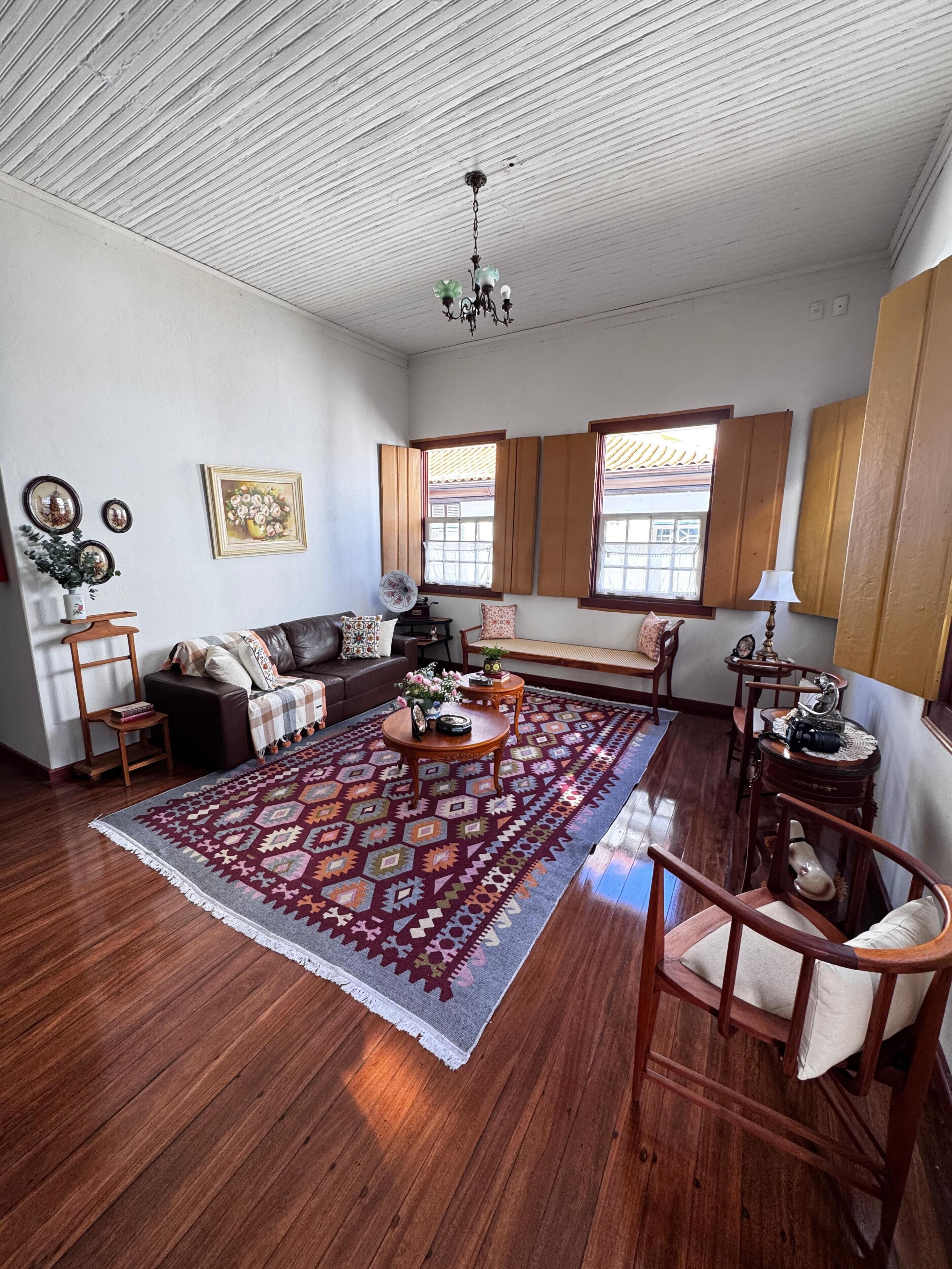 A spacious living room with a large wooden floor, a patterned rug, a sofa, a coffee table, and a wooden chair. There are two windows with wooden shutters, a ceiling fan, and a chandelier.