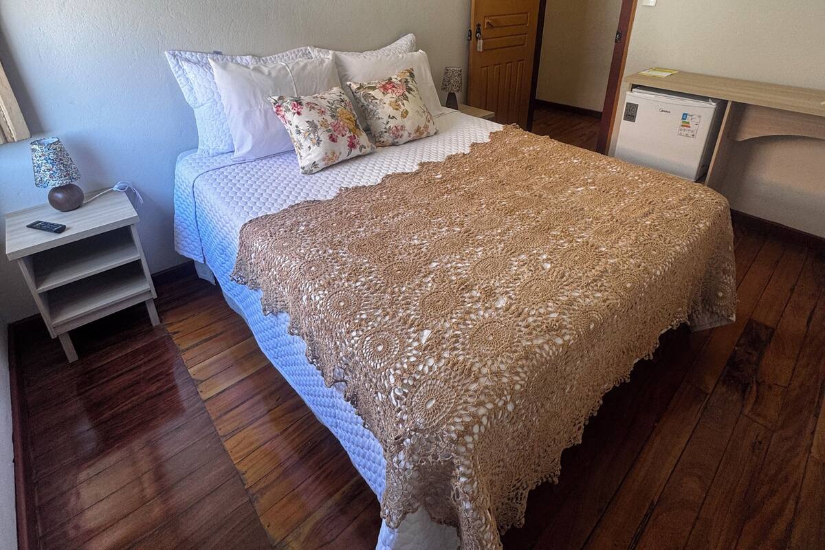 Un dormitorio con piso de madera, una cama con colcha blanca y almohadas florales, una mesita de noche de madera con un control remoto, y una puerta con espejo.
