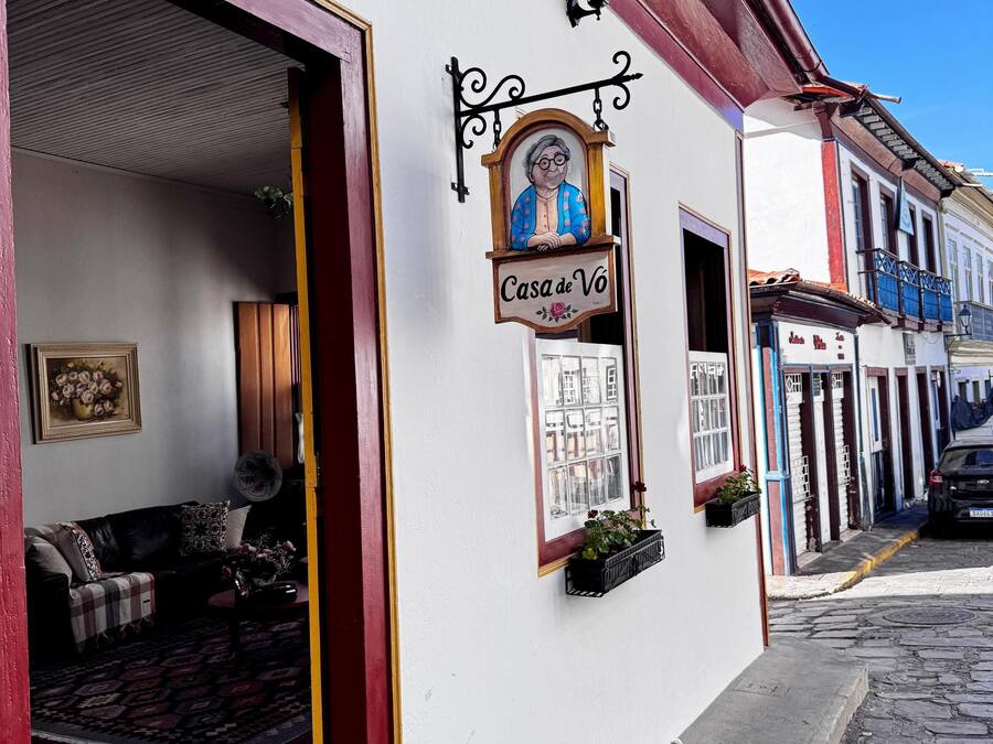 Vista da rua com um edifício identificado como 'Casa de Vó', com fachada tradicional, entrada visível e um pequeno espaço ao ar livre com um ventilador e almofadas.