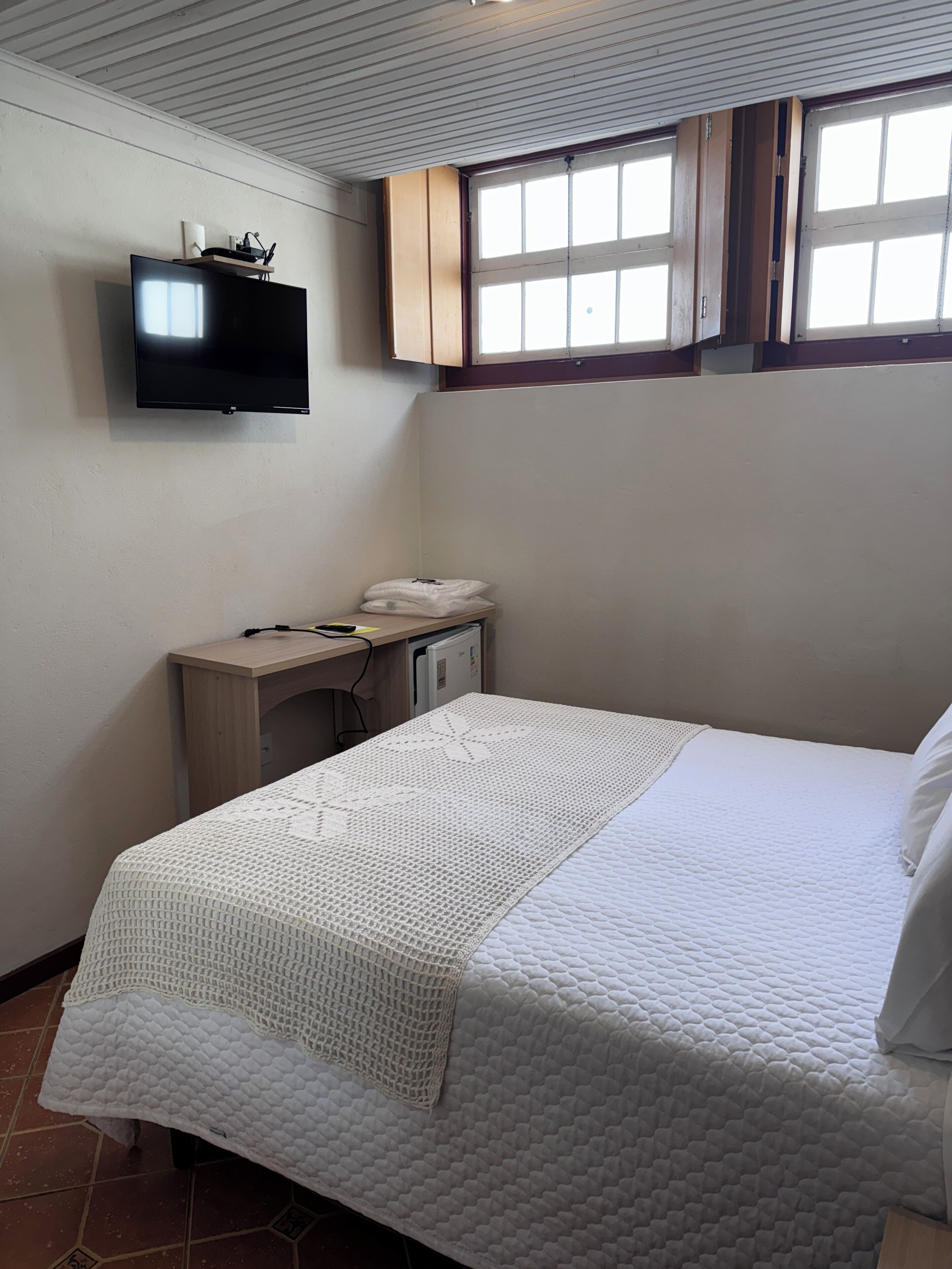 A bedroom with a bed, a desk with a computer, a TV mounted on the wall, and a window with wooden shutters.