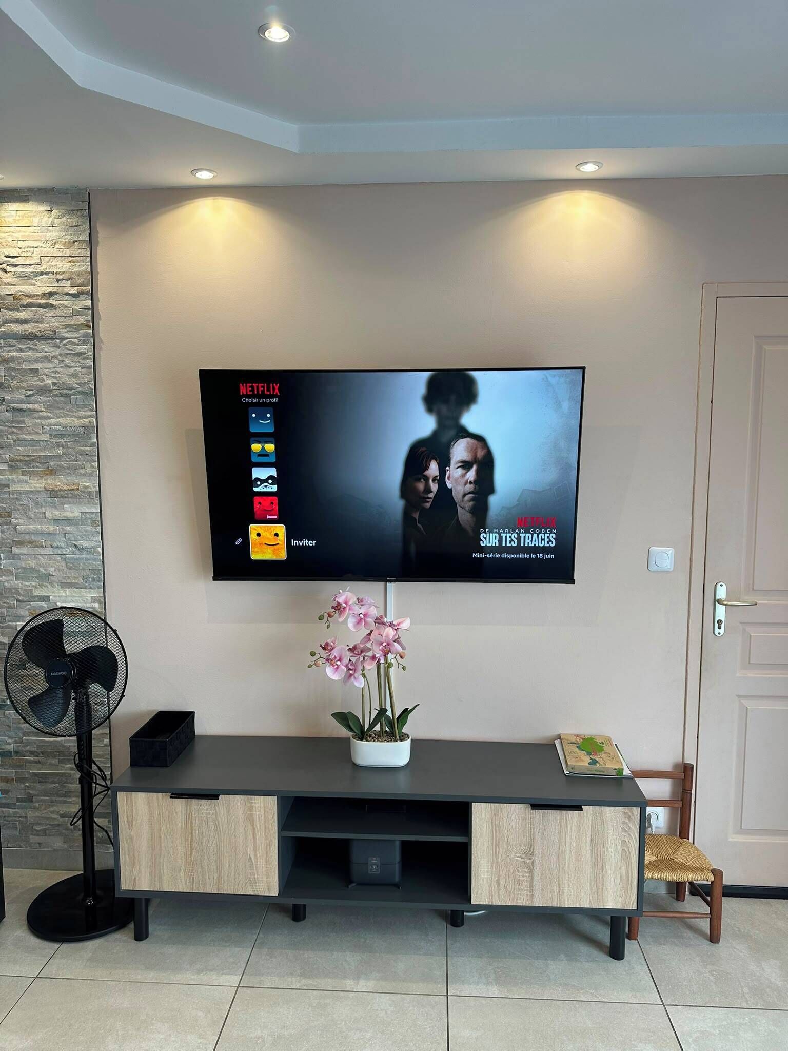 A modern living room with a flat-screen TV mounted on the wall, a wooden console table with a potted orchid, and a standing fan.