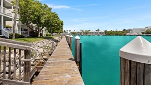 Une jetée en bois mène à une étendue d’eau turquoise, avec une rangée de bâtiments blancs sur la gauche et un ciel bleu clair au-dessus.