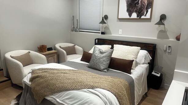 A bedroom with a bed, two chairs, a nightstand, a window with blinds, a wall-mounted lamp, a framed picture, and a scale on the floor.