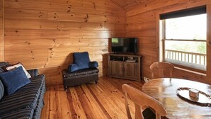 A cozy cabin interior with a wooden floor, a sofa, a leather armchair, a television on a wooden cabinet, and a dining table with chairs. - Cabin #4 The Sky Retreat - Off the Blue Ridge Parkway (Purlear)