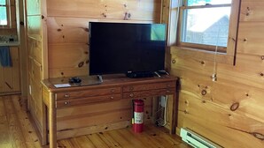 Ein Zimmer in einer Holzhütte mit einem Fernseher auf einem Holztisch, einem Fenster mit Jalousien und einem Bett mit gestreifter Bettwäsche.