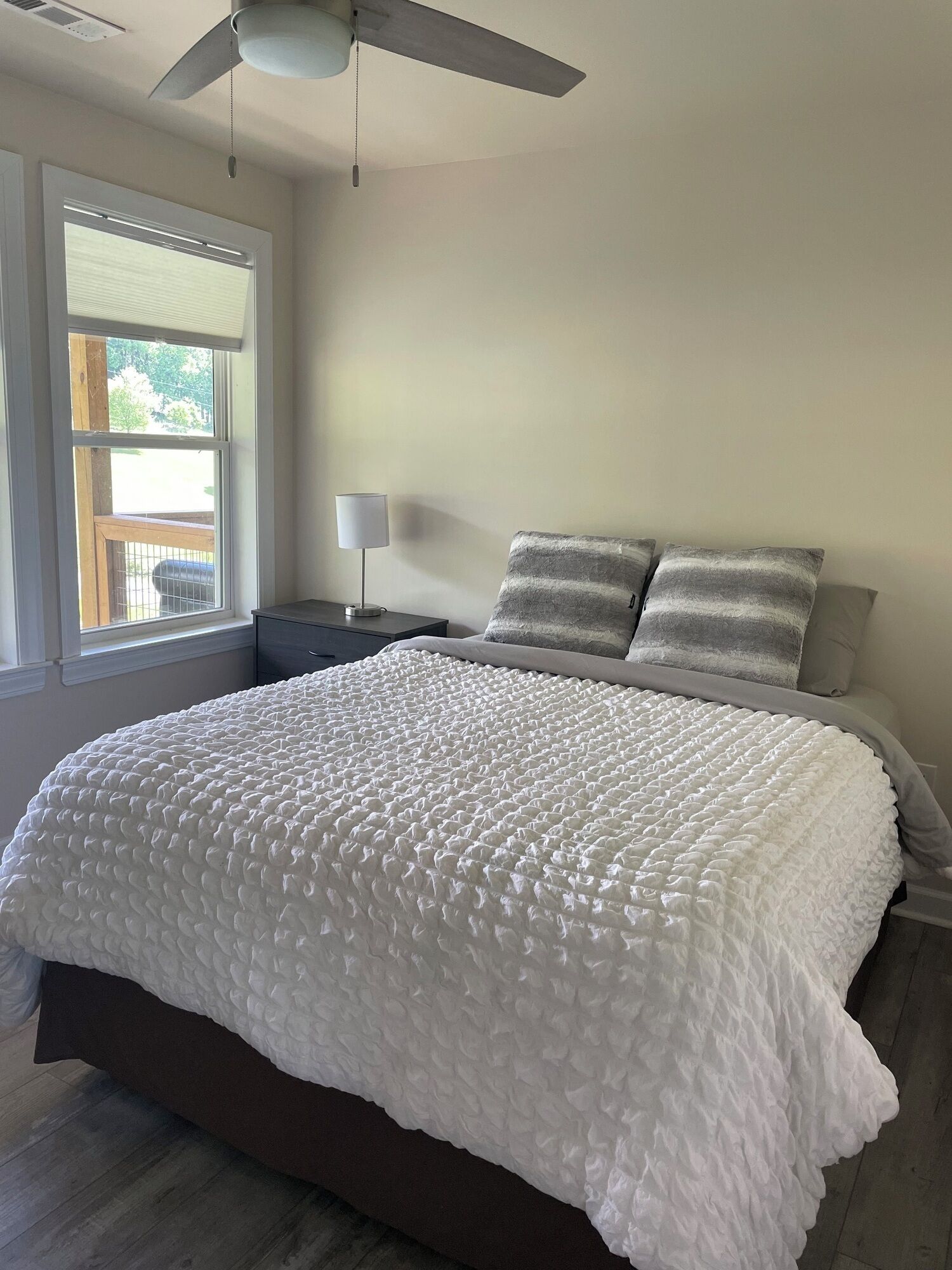 A bedroom with a large bed, a ceiling fan, a nightstand with a lamp, and a window with blinds.