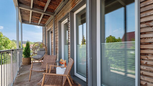 A wooden deck with wicker chairs and a small table, large windows with blinds, and a view of greenery. - Novita Apartment - balcony | free parking | central (Bad Harzburg)