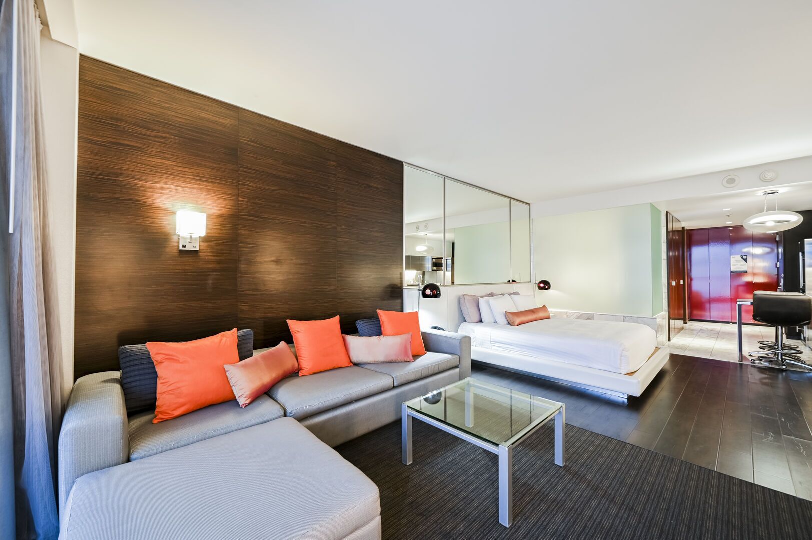 A modern hotel room with a grey sofa, a glass coffee table, and a bed with white linens.