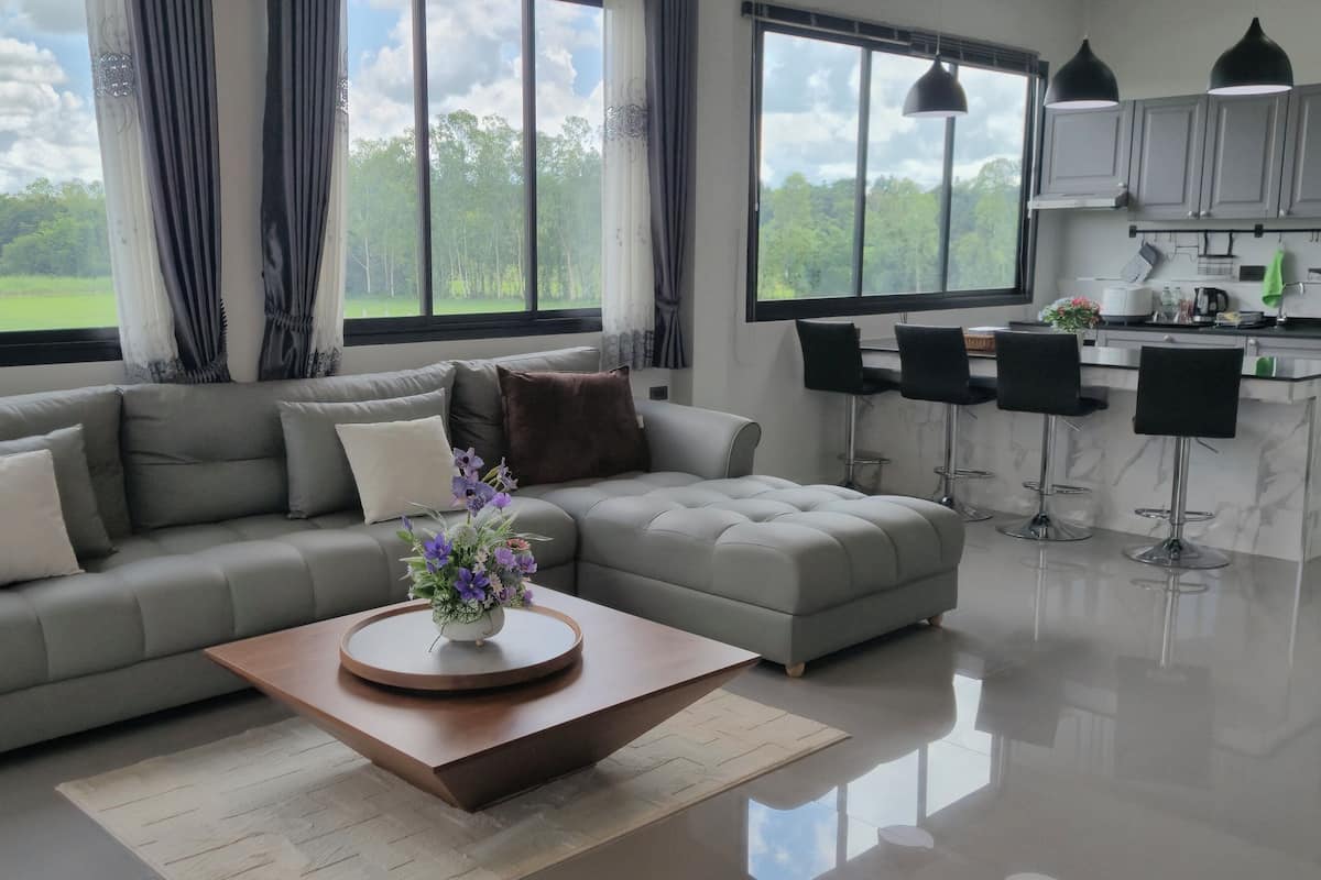 Una sala de estar moderna con un sofá seccional gris, una mesa redonda de centro con un jarrón de flores y vistas a un paisaje verde a través de grandes ventanales.