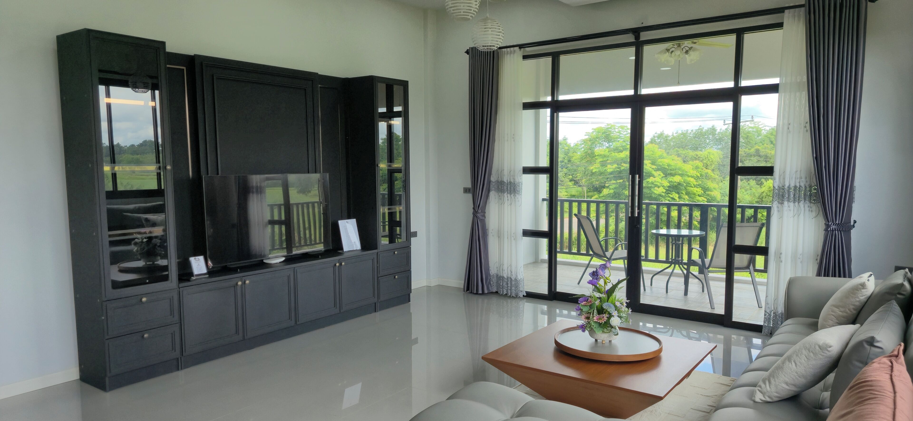 A modern living room with a sofa, a coffee table, and a TV on a cabinet. There is a balcony with a view of greenery.