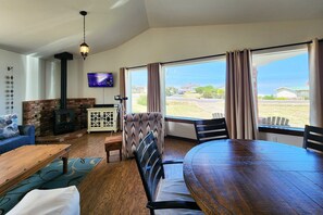 A cozy living room with a fireplace, a TV, a dining table, and large windows offering a view of the outdoors. - Ocean View, Walk to Everything! Dog Friendly! Cute Yachats, Oregon Home! (Yachats)