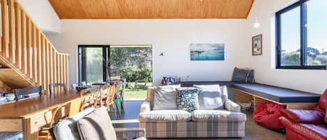 A spacious living room with a wooden ceiling, a dining area with a wooden table and chairs, a sofa with patterned cushions, a red leather couch, and a large window offering a view of the outdoors.