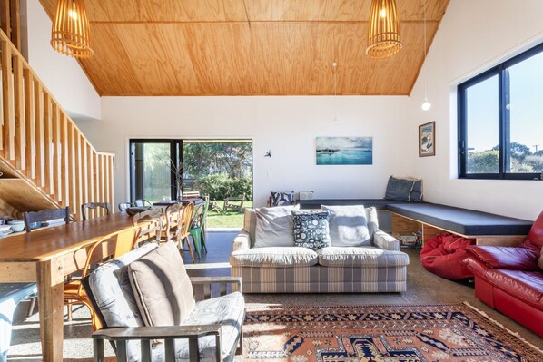 A spacious living room with a wooden ceiling, a dining area with a wooden table and chairs, a sofa with patterned cushions, a red leather couch, and a large window offering a view of the outdoors.