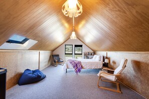 Un dormitorio acogedor en el ático, con techo inclinado, una cama con manta morada, una mecedora de madera y un sillón de peluche azul.