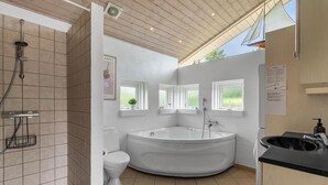 Un baño moderno con una bañera independiente, inodoro y ducha. Hay ventanas que dejan entrar luz natural y ofrecen vistas a la vegetación.