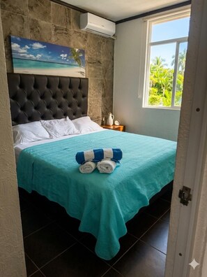 A hotel room with a bed, a painting of a beach, a window with a view of greenery, and a door.