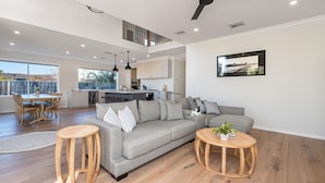 Amplia sala de estar con un sofá gris, una mesa de centro de madera y una mesa de comedor con sillas. Se ve una cocina al fondo, con vistas al exterior.