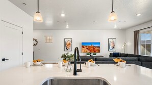 Een moderne keuken met een roestvrijstalen spoelbak, een eethoek met een tafel en stoelen, een grijze bank en een aan de muur bevestigde televisie.