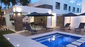 Un área de piscina moderna al aire libre con sillones de descanso y sombrillas, rodeada por un edificio con grandes ventanales.