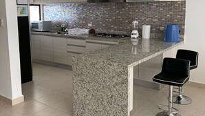 Una cocina moderna con encimeras de granito, microondas integrado, estufa a gas y cafetera. Hay dos taburetes negros y un bote de basura azul.