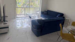 Una sala moderna con un sofá azul, una mesa de centro de vidrio, un sillón amarillo y un televisor sobre un mueble.