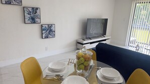Un comedor con mesa de vidrio, sillas amarillas y un frutero. Se ve un sofá y un televisor al fondo.